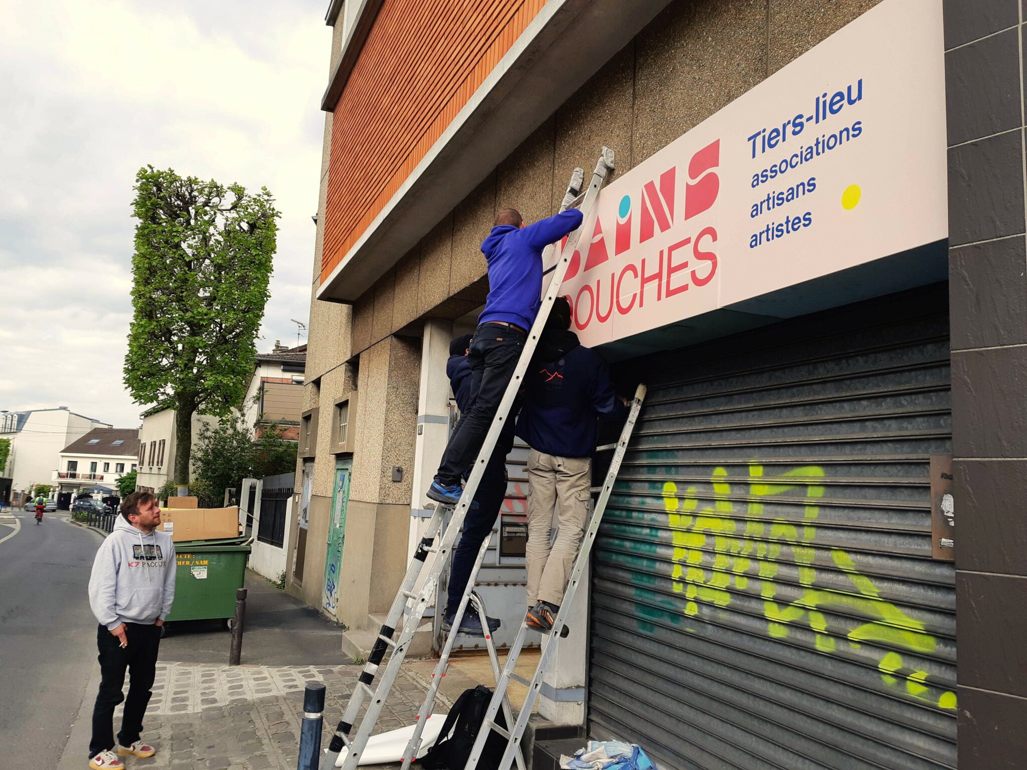 Quatre personnes, trois échelles, pour installer une signalétique des Bains Douches sur le bâtiment